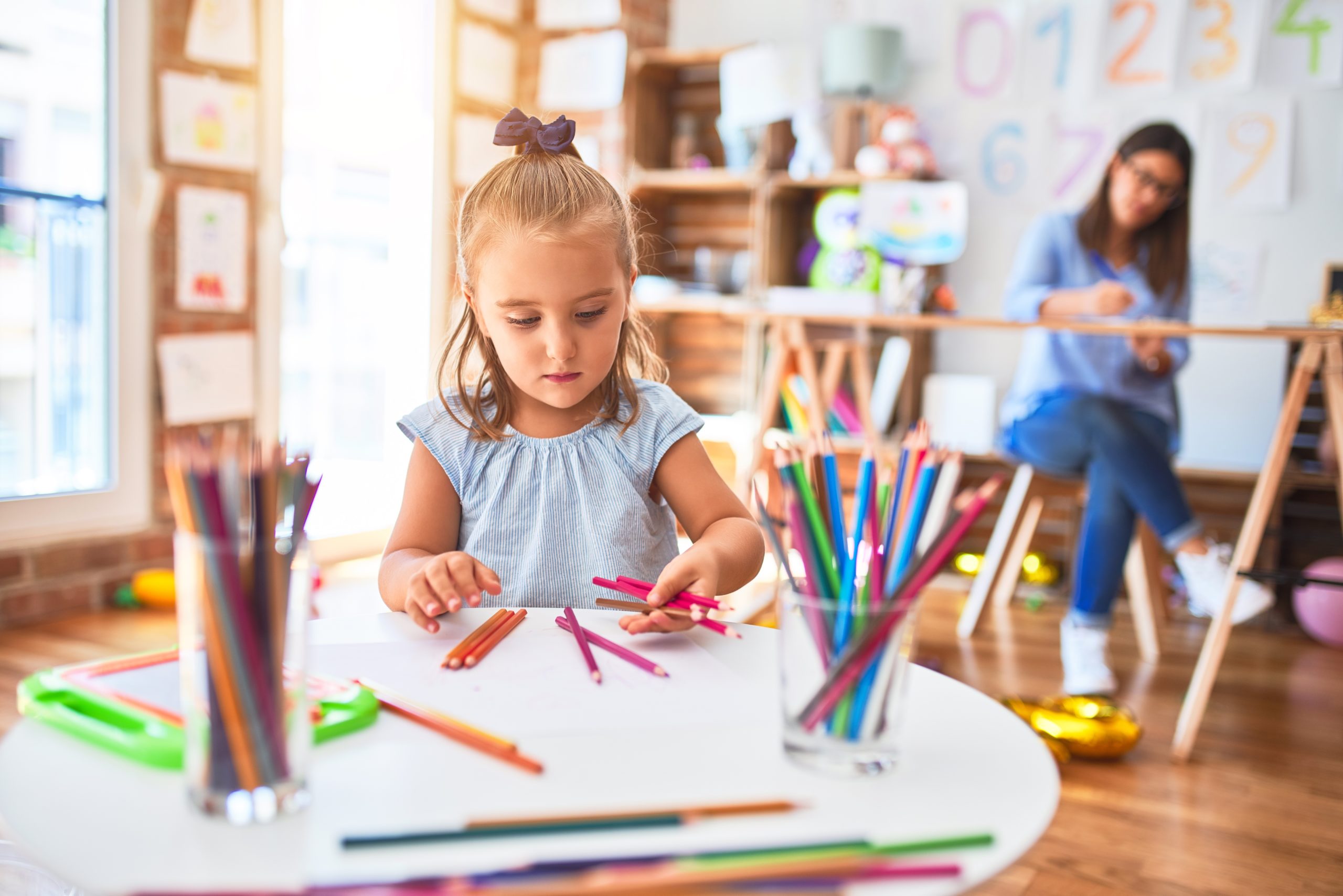 Caucasian girl kid playing and learning at playschool with female teacher. Mother and daughter at playroom around toys drawing with pencil colors